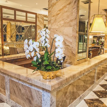 Windsor Marapendi Hotel Rio de Janeiro Eksteriør billede The photo depicts a luxurious hotel lobby featuring a polished marble reception desk. There are elegant white orchids arranged in a gold pot on the counter, adding a touch of sophistication. The surrounding interior showcases marble walls, stylish li
