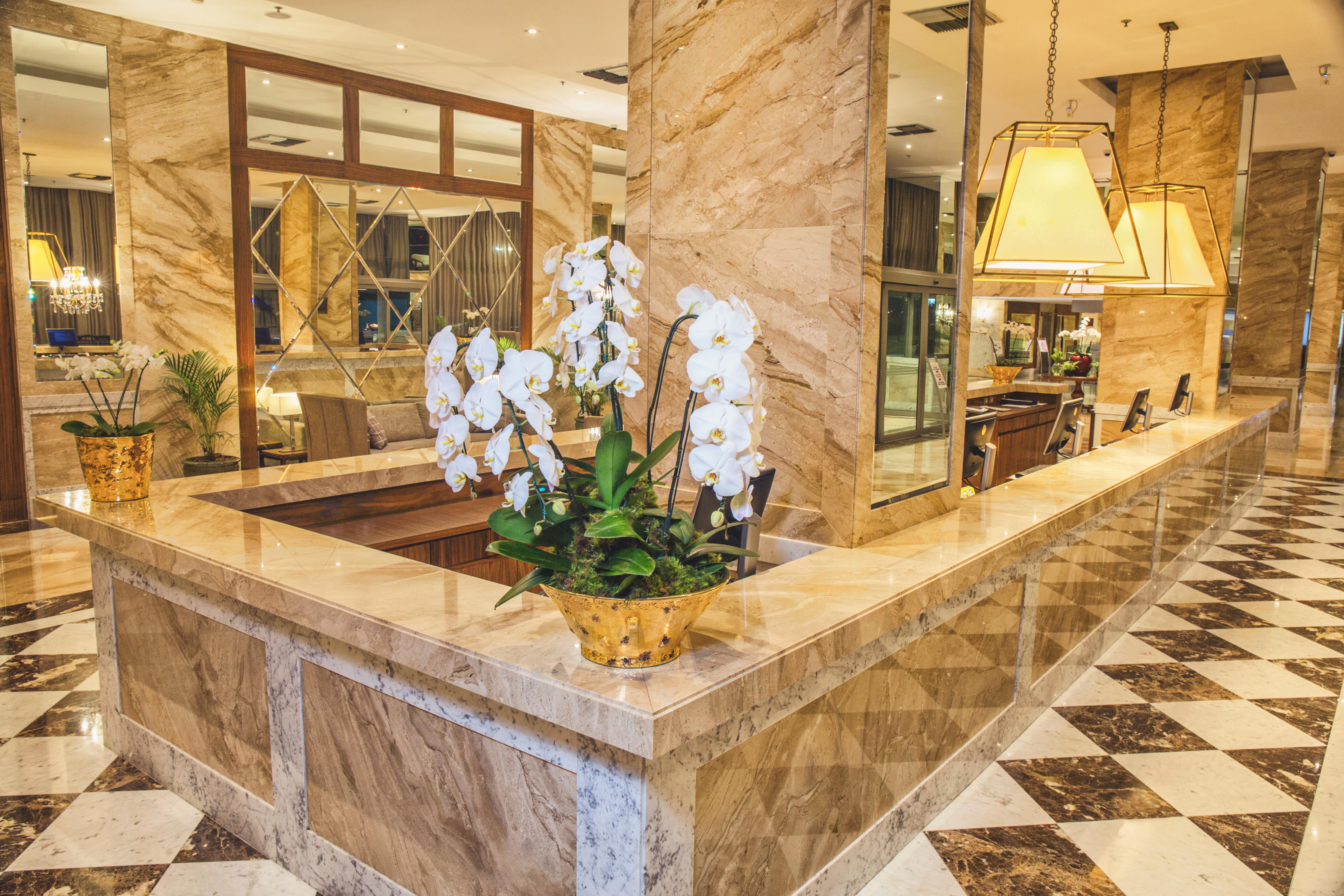 Windsor Marapendi Hotel Rio de Janeiro Eksteriør billede The photo depicts a luxurious hotel lobby featuring a polished marble reception desk. There are elegant white orchids arranged in a gold pot on the counter, adding a touch of sophistication. The surrounding interior showcases marble walls, stylish li