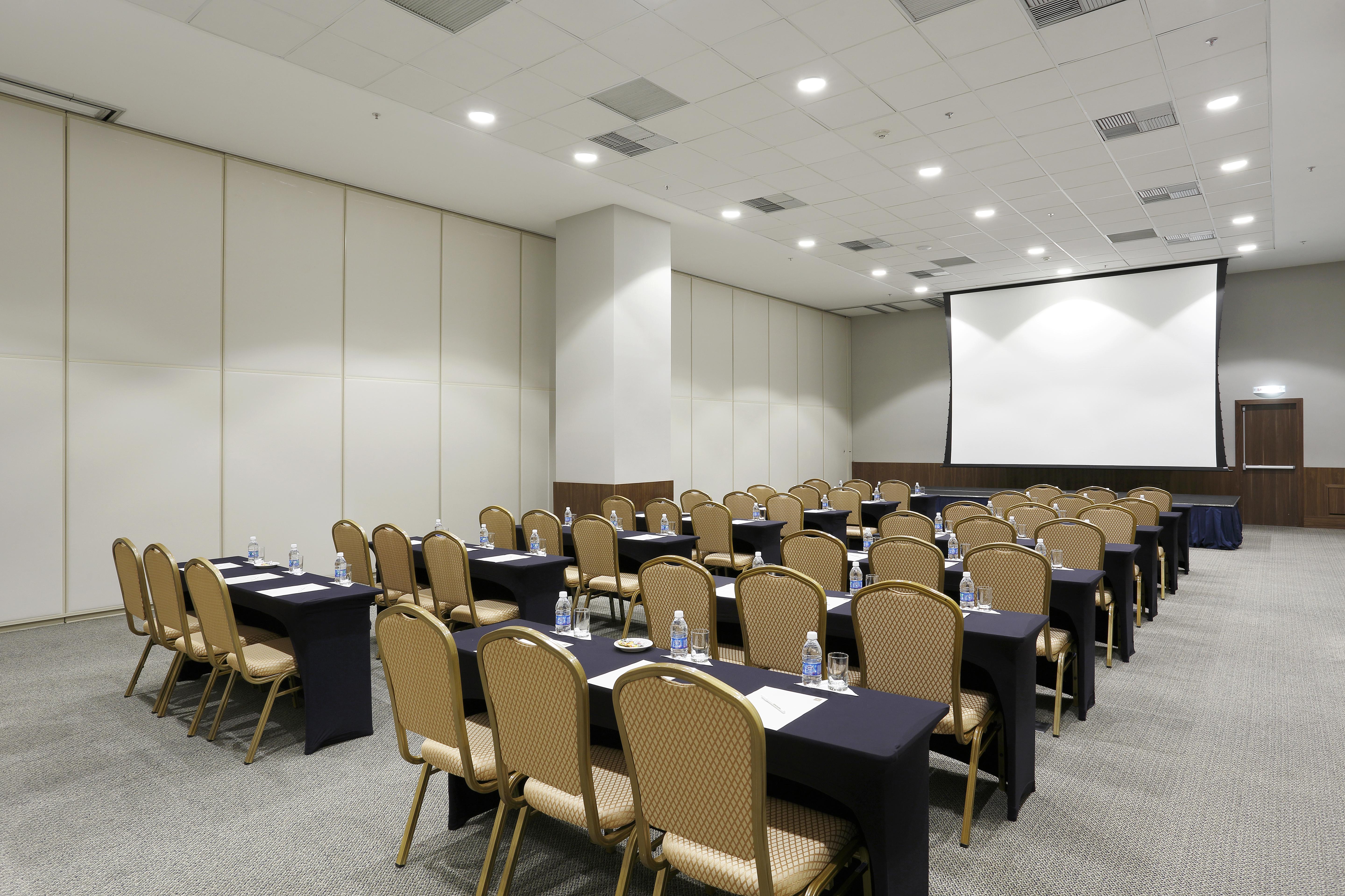 Windsor Marapendi Hotel Rio de Janeiro Eksteriør billede The photo shows a conference or meeting room arranged with several rows of tables and chairs. The tables are covered with dark tablecloths and have water bottles placed on them. The room is bright and spacious, featuring a large projection screen on 